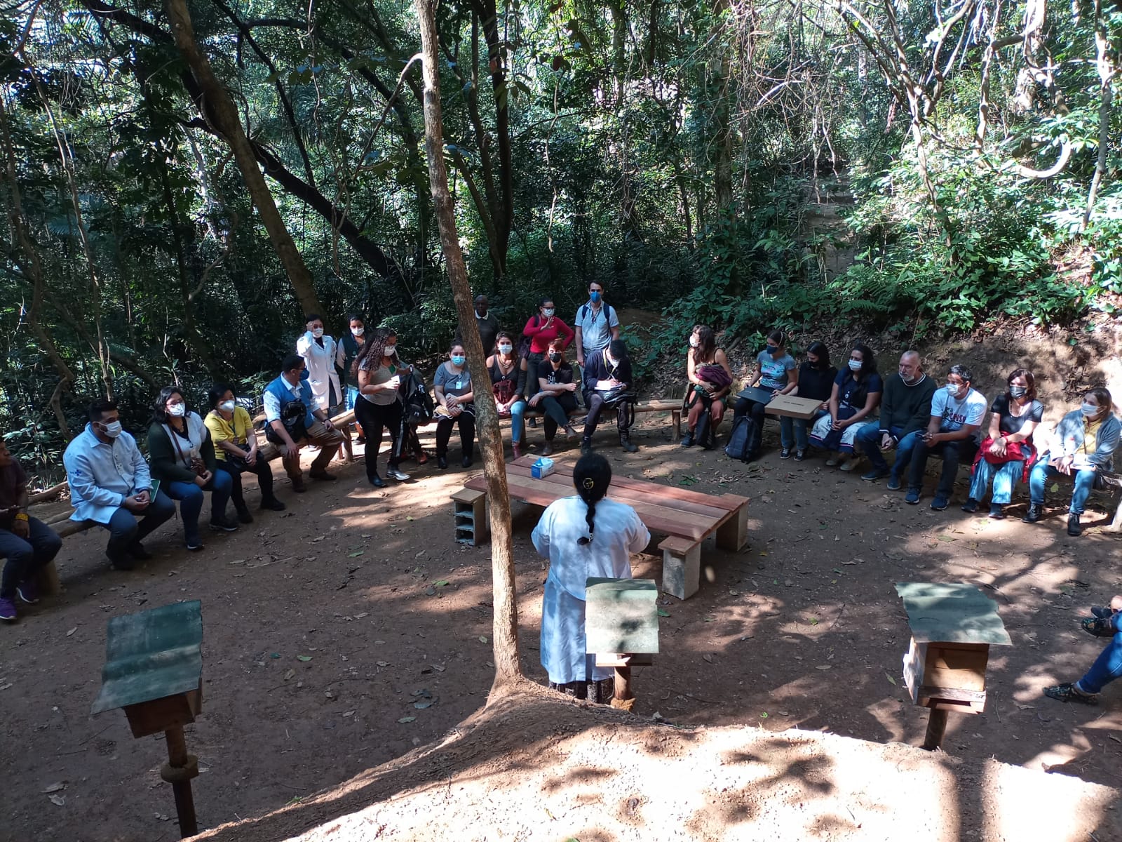 A foto mostra uma mulher de cabelos compridos e escuros, usando um jaleco branco; ela está de costas para o fotógrafo, falando para um grupo de cerca de 20 pessoas, homens e mulheres, sentados em semicírculo em uma clareira no meio da mata